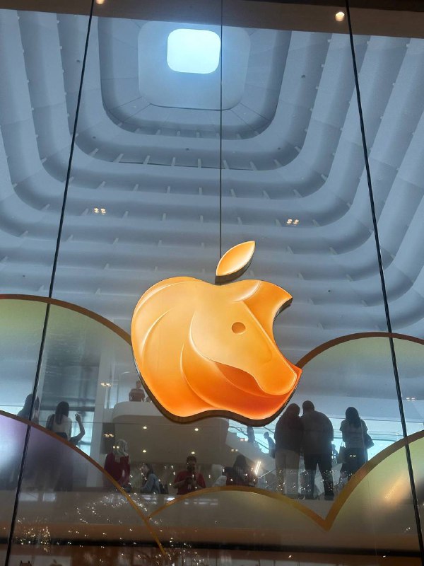 Крупный план логотипа Apple в витрине торгового центра с отражением людей внутри — минималистичный городской кадр, использованный как визуал.