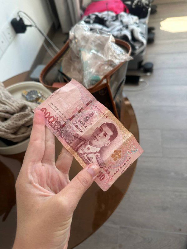 Крупный план руки, держащей купюру тайского бата на фоне сумки и вещей — визуальный элемент, ассоциирующийся с деньгами и путешествиями.