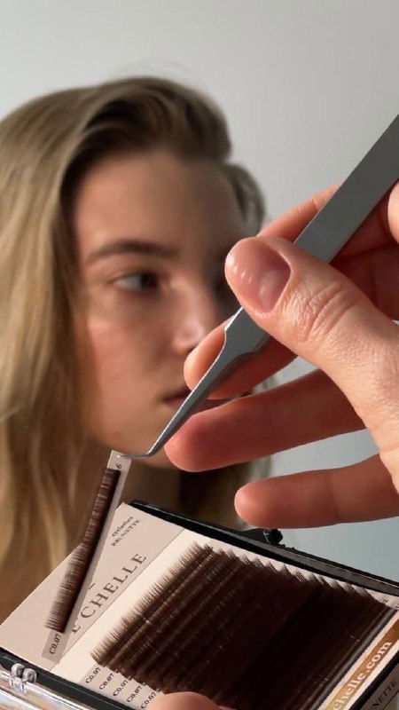 Крупный план рук мастера с пинцетом над палеткой ресниц и размытым лицом модели на заднем плане — визуал процесса работы в студии.