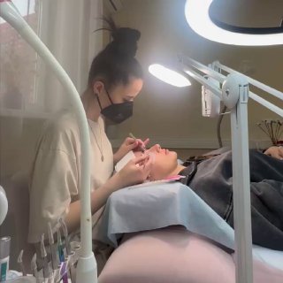 Лэшмастер в маске наносит наращивание ресниц клиентке, пациентка лежит под кольцевой лампой в салоне с инструментами рядом.