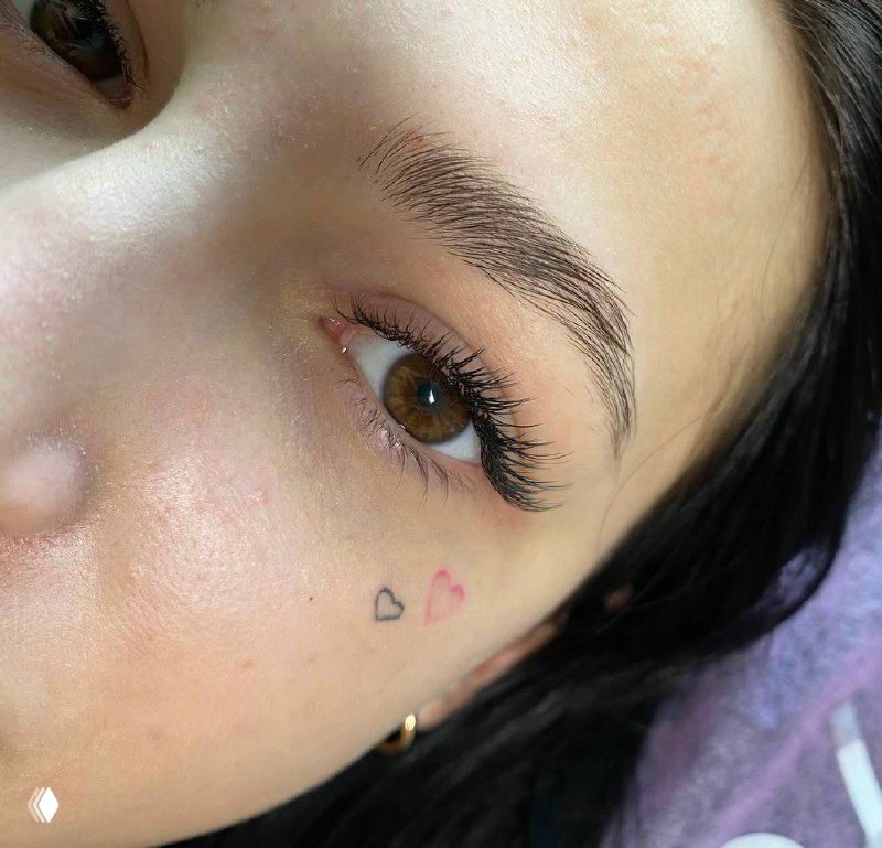 Крупный план глаза с наращиванием ресниц и маленькой тату‑деталью у скулы; тёплый тон кожи, аккуратный объём и естественное моделирование.