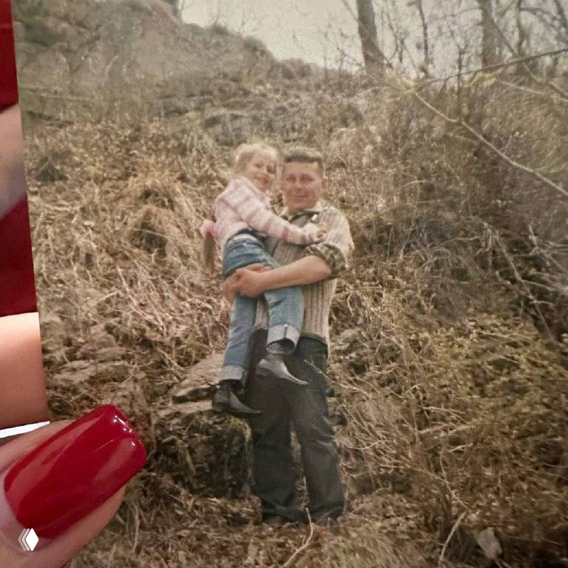Архивное фото: отец держит маленькую девочку на руках на фоне природы, снимок подчёркивает сельское детство и семейные узы.