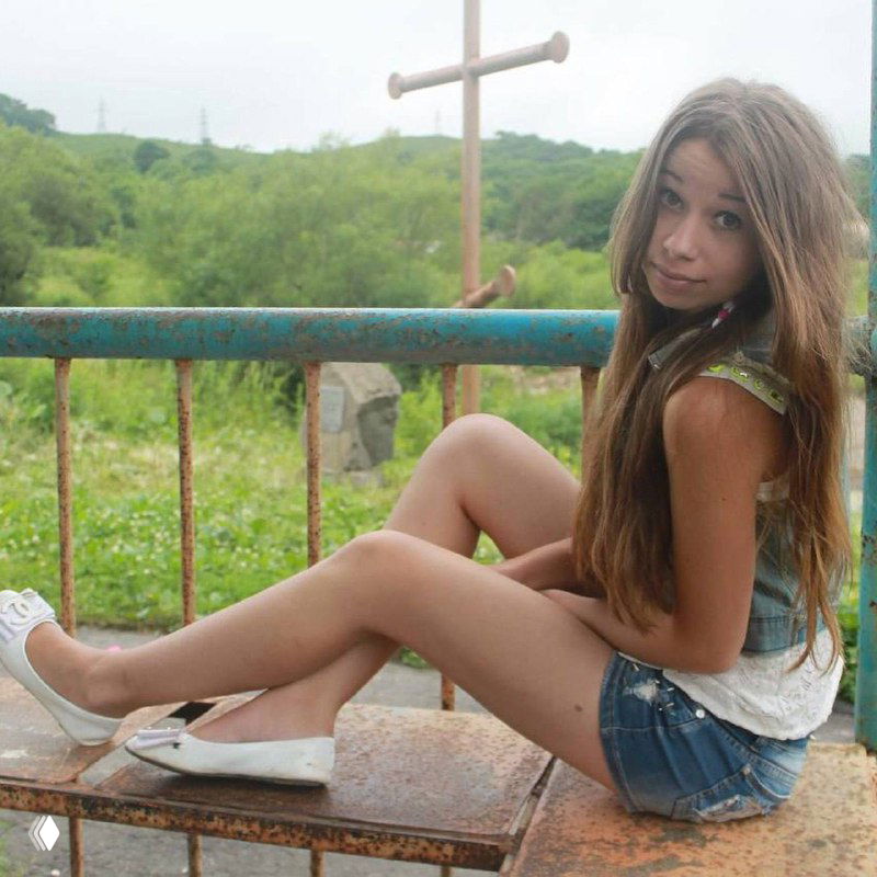 Подростковый снимок девушки на металлическом ограждении с крестом и зеленью на заднем плане, показывающий юношеские годы в посёлке до переезда.