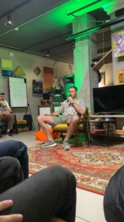 Спикер в шортах и футболке сидит с микрофоном в неформальном помещении; видны ковёр, экран и часть аудитории.