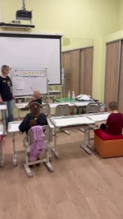 Учебный класс с доской, преподавателем и несколькими детьми за партами; один ребёнок сосредоточенно сидит и решает задачу в процессе занятия.