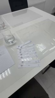 На белом столе бумажные тест‑полоски для парфюма, рядом блокнот и стакан воды, видны отражения ламп — кадр с мастер‑класса по ароматам.