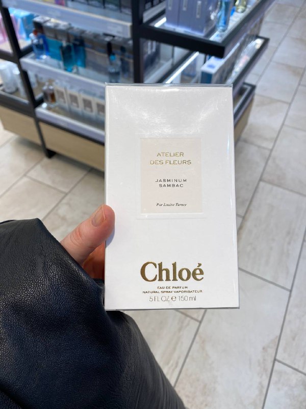 Крупный план коробки духов Chloé в руке на фоне полок дьюти‑фри; упаковка белая с золотым тиснением, магазинный интерьер виден позади.