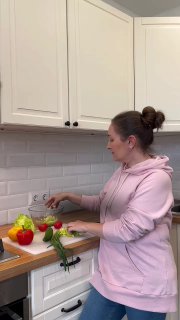 Женщина в розовом худи нарезает овощи на разделочной доске на фоне светлой кухни; на столе видны перцы и зелень, вид сбоку.