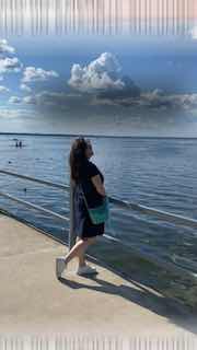 Женщина в тёмном платье с сумкой стоит у перил на набережной, смотрит на широкую водную гладь и облачное небо; летний городской пейзаж.