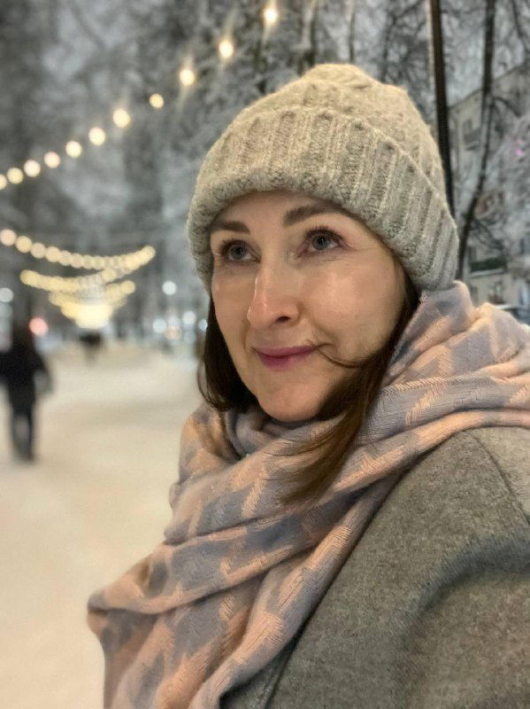 Портрет женщины в тёплой шапке и шарфе на фоне зимней аллеи с размытыми гирляндами; кадр снят крупным планом, тёплый свет.