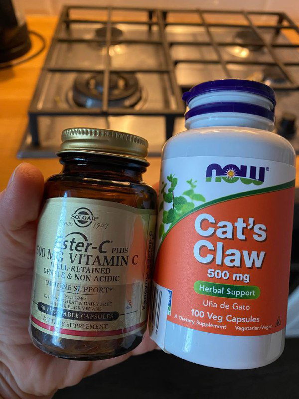 Две банки пищевых добавок на фоне кухонной плиты: витамин C и бутылочка Cat's Claw; крупный план этикеток и крышек.