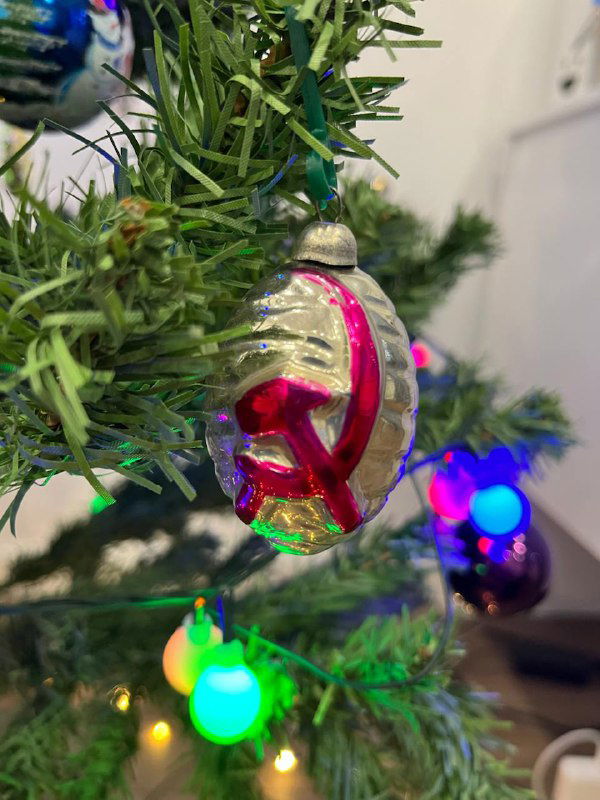 Крупный стеклянный ёлочный шар-игрушка с яркой красной деталью на ветке искусственной ёлки, размытый фон домашнего интерьера.