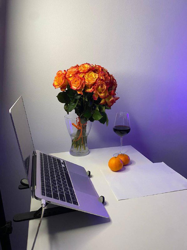 Рабочее место с ноутбуком на подставке, букетом роз, бокалом вина и мандаринами — праздничный рабочий уголок с мягкой подсветкой.