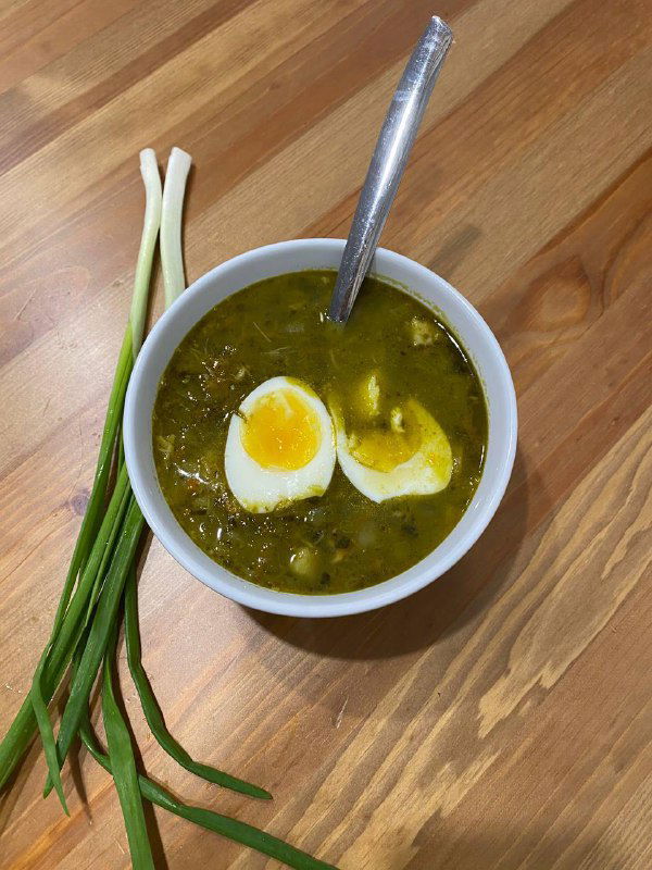 Порция тёплого зелёного супа в миске с половинками варёного яйца и зелёным луком, вид сверху на деревянном столе в домашней обстановке.