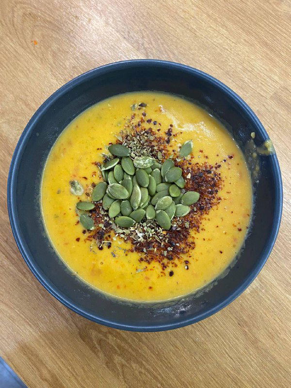 Крем‑суп из тыквы в синей миске, украшенный семечками тыквы и пряными хлопьями, вид сверху на кухонном столе.