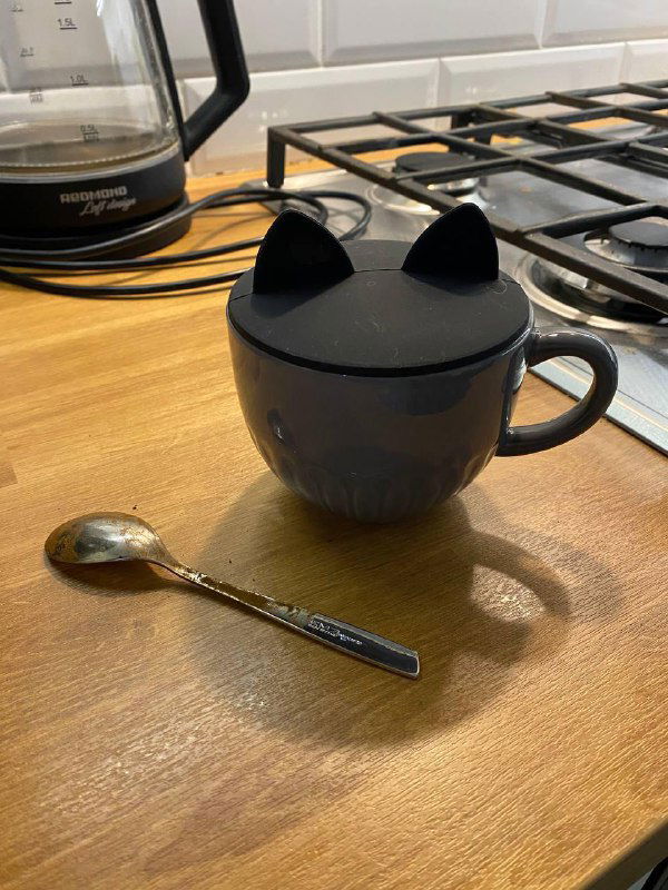 Кружка с крышкой в форме ушек, стоящая на кухонной стойке рядом со столовыми приборами, создаёт уютную домашнюю атмосферу.