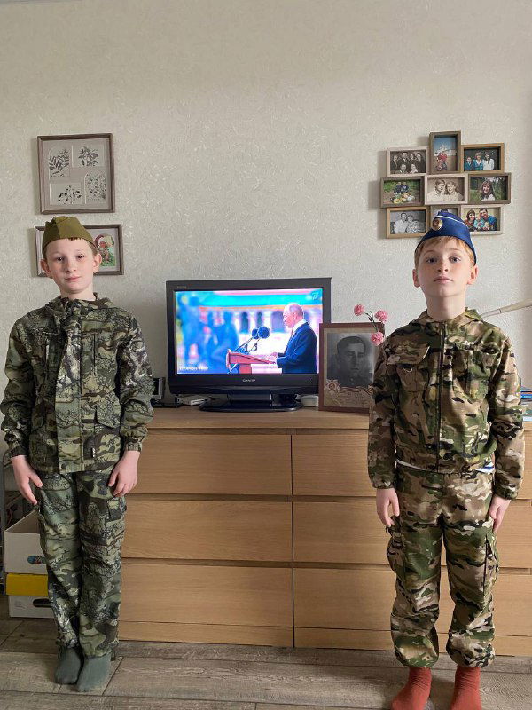 Двое мальчиков в камуфляже стоят у комода перед телевизором, где идёт парад; праздничный семейный интерьер и военная атрибутика.