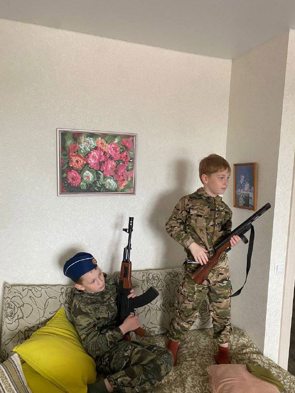 Мальчики в камуфляже с имитационными винтовками позируют в гостиной рядом с диваном и предметами декора — кадр памяти к празднику.