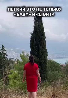 Женщина в ярко‑красном платье идёт по тропинке к высокому кипарису на фоне моря и облачного неба, вид сзади.