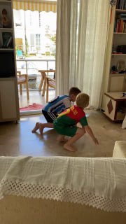Двое детей в гостиной выполняют упражнения на полу: игровой домашний контекст, дети в движении рядом с мебелью и открытым балконом.