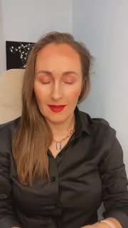 Портрет женщины в интерьере: сидит перед камерой с закрытыми глазами, в тёмной блузе и украшении на шее, нейтральный фон