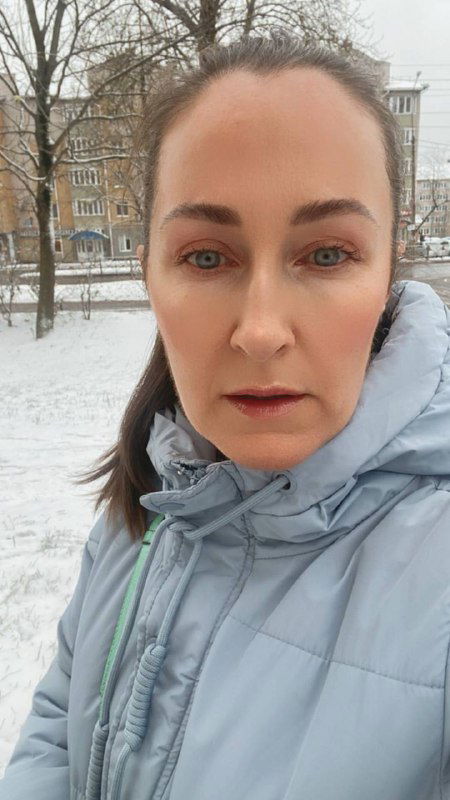 Крупный уличный портрет автора зимой в светлой куртке; на заднем плане снег, деревья и жилой дом, выражение задумчивое и спокойное.