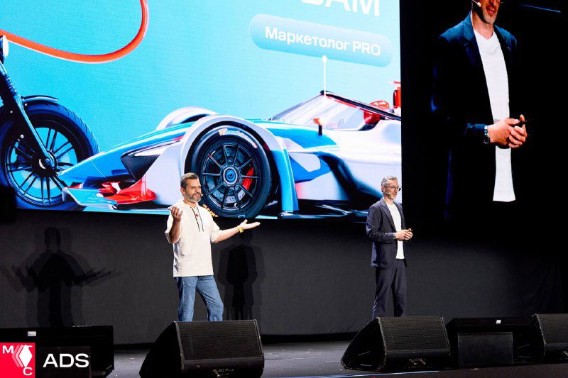 Два спикера на сцене у большого экрана с изображением гоночного автомобиля: динамичная подача, микрофоны и зрители внизу.