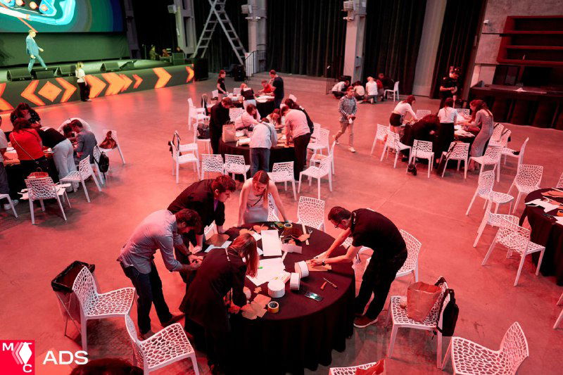 Участники за круглым столом в зоне воркшопа: команды обсуждают задачи, ноутбуки и материалы на столах, вид сверху.