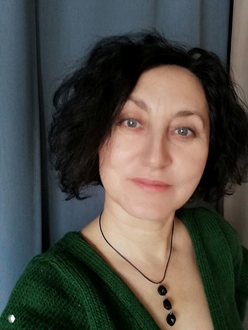 Портрет женщины среднего возраста в зелёном свитере, снятый крупным планом на фоне штор; нейтральное выражение лица и мягкое освещение.