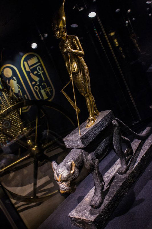 Боковой крупный план статуи фараона/божества и леопарда на постаменте в тёмной витрине — видны детали украшений и элементы композиции.