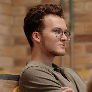 Крупный план молодого мужчины в очках на конференции, внимательный взгляд, интерьер конференц-зала с кирпичным фоном на заднем плане