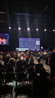 Аудитория на конференции TG Conf: участники и пустые стулья, вид на сцену и крупный экран, где показывают слайды презентаций.