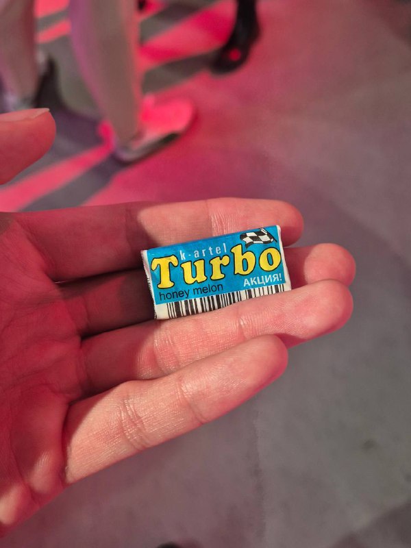 Фотография руки, держащей жвачку Turbo — предмет раздатки на мероприятии, крупный план упаковки и пола конференц-зоны в фоне.