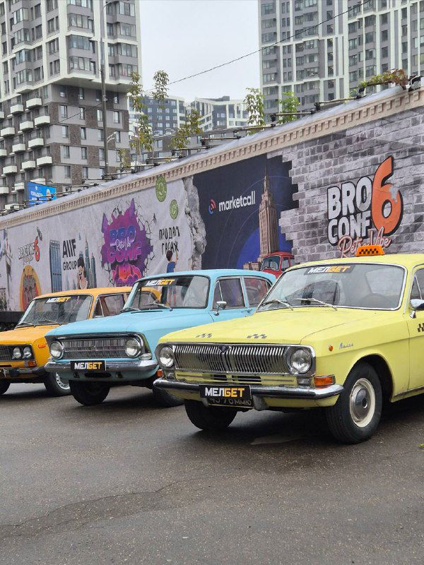 Ретро‑автомобили на фоне уличного мурала с рекламой BROCONF-6 — городской кадр рядом с зоной конференции, несколько машин в ряд.