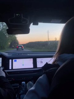 Вид из салона автомобиля на дорогу и приборную панель; пассажирка и водитель на фоне вечерней трассы и закатного неба.