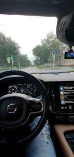 Вид из салона автомобиля с места водителя: рулевое колесо, приборная панель и центральная консоль на фоне загородной дороги, снято вечером с освещённой фарами трассой.