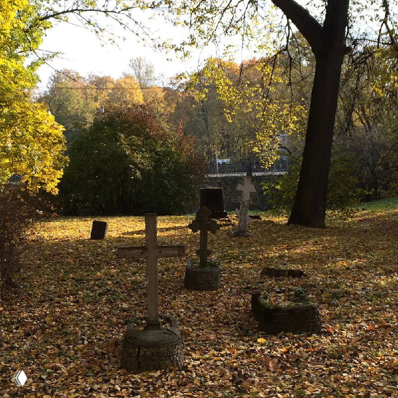 Несколько деревянных и каменных крестов на кладбище в осеннем парке; желтые листья устилают землю вокруг надгробий и деревья на фоне.