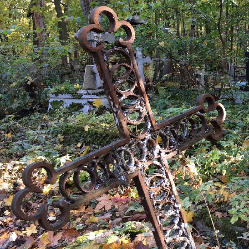 Ржавый кованый крест, наклонённый на старом кладбище среди зарослей и опавших листьев; сочетаются текстуры металла, мха и листвы.