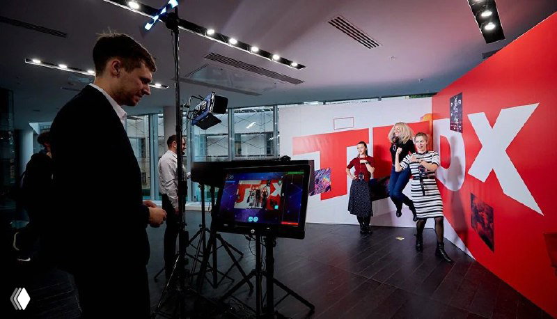 Интерьерная фотозона с красной брендированной стеной и большой буквой TRIX, сотрудники и гости взаимодействуют с экраном и техникой для съёмки.