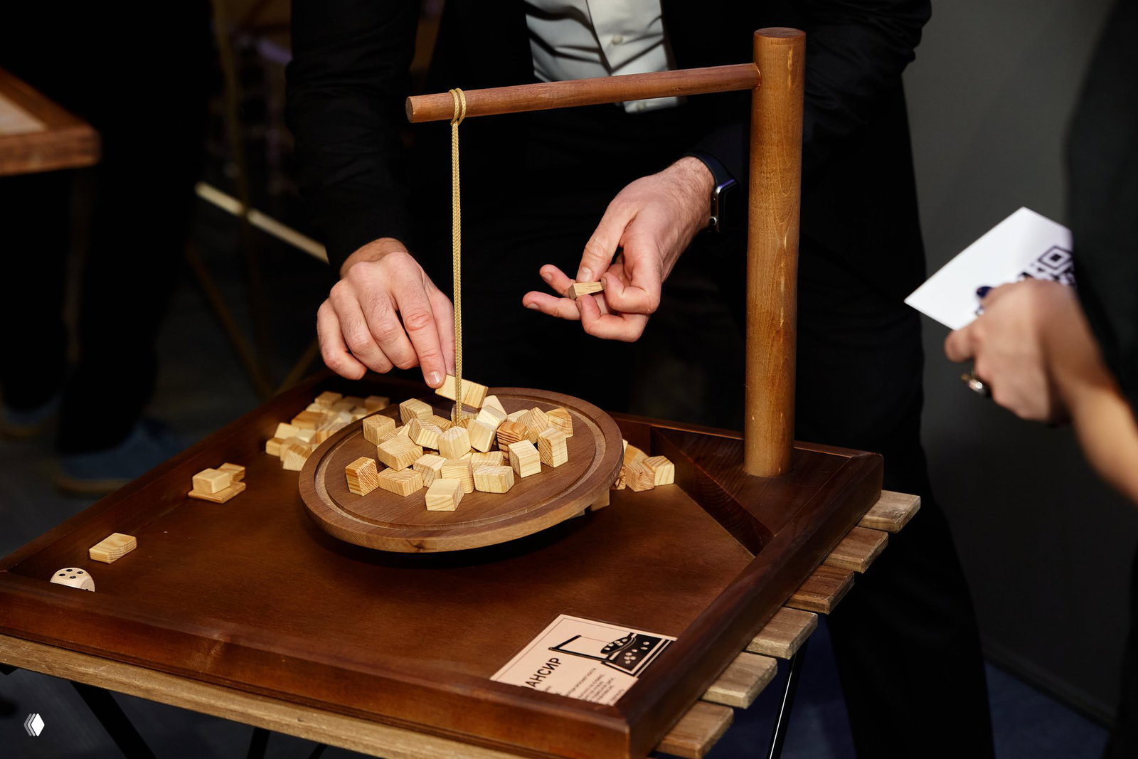 Руки ведущего расставляют деревянные кубики на столе с подвешенным элементом игры, видна табличка с информацией и участники в кадре.