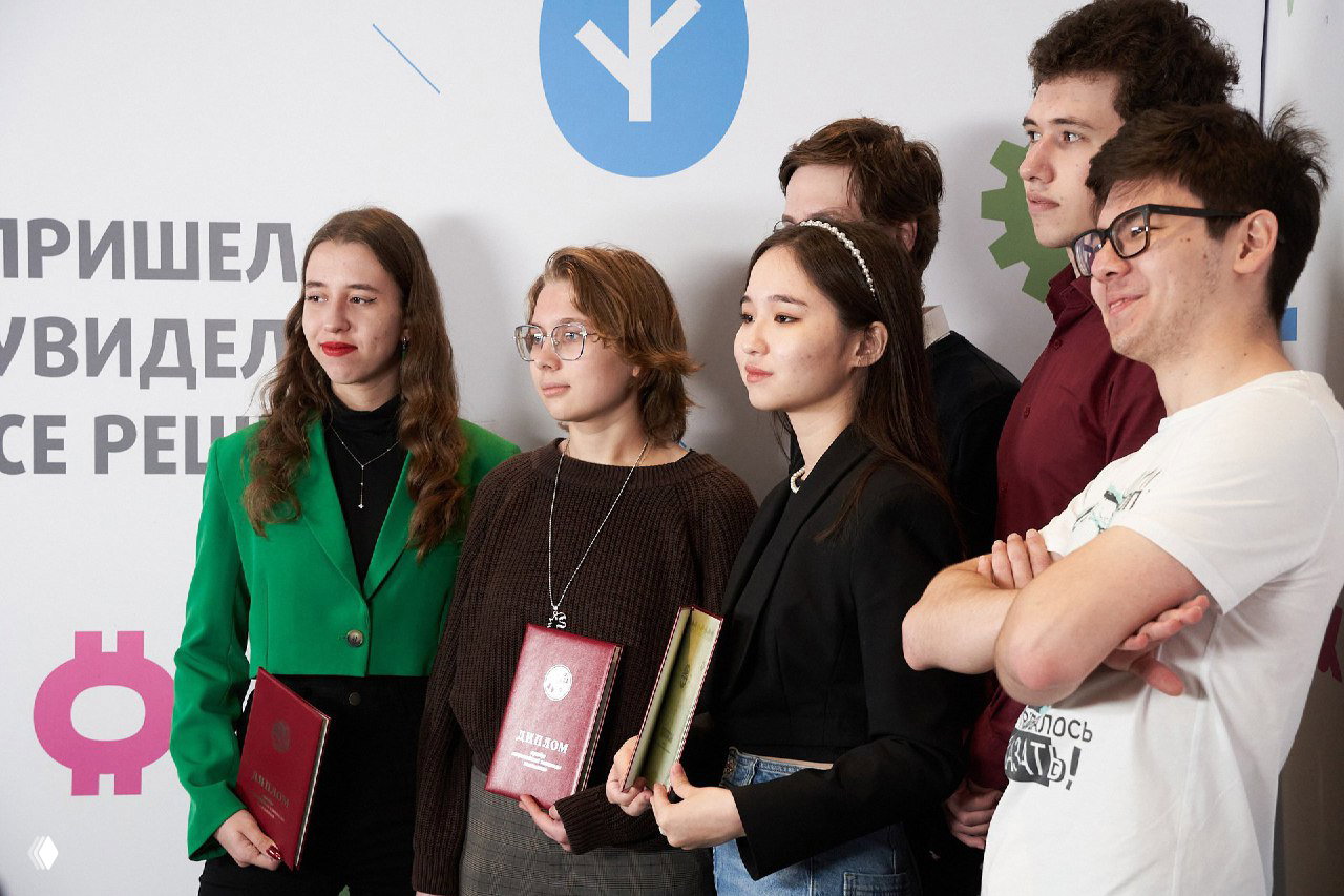 Группа школьников и студентов с дипломами позирует на фоне брендированного пресс‑волла; кадр иллюстрирует победителей Всероссийской олимпиады на мероприятии.