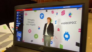 Экран монитора управления фотокабины показывает кадр с участницей на фоне пресс‑волла; виден интерфейс съемки и экран превью результата для гостей.