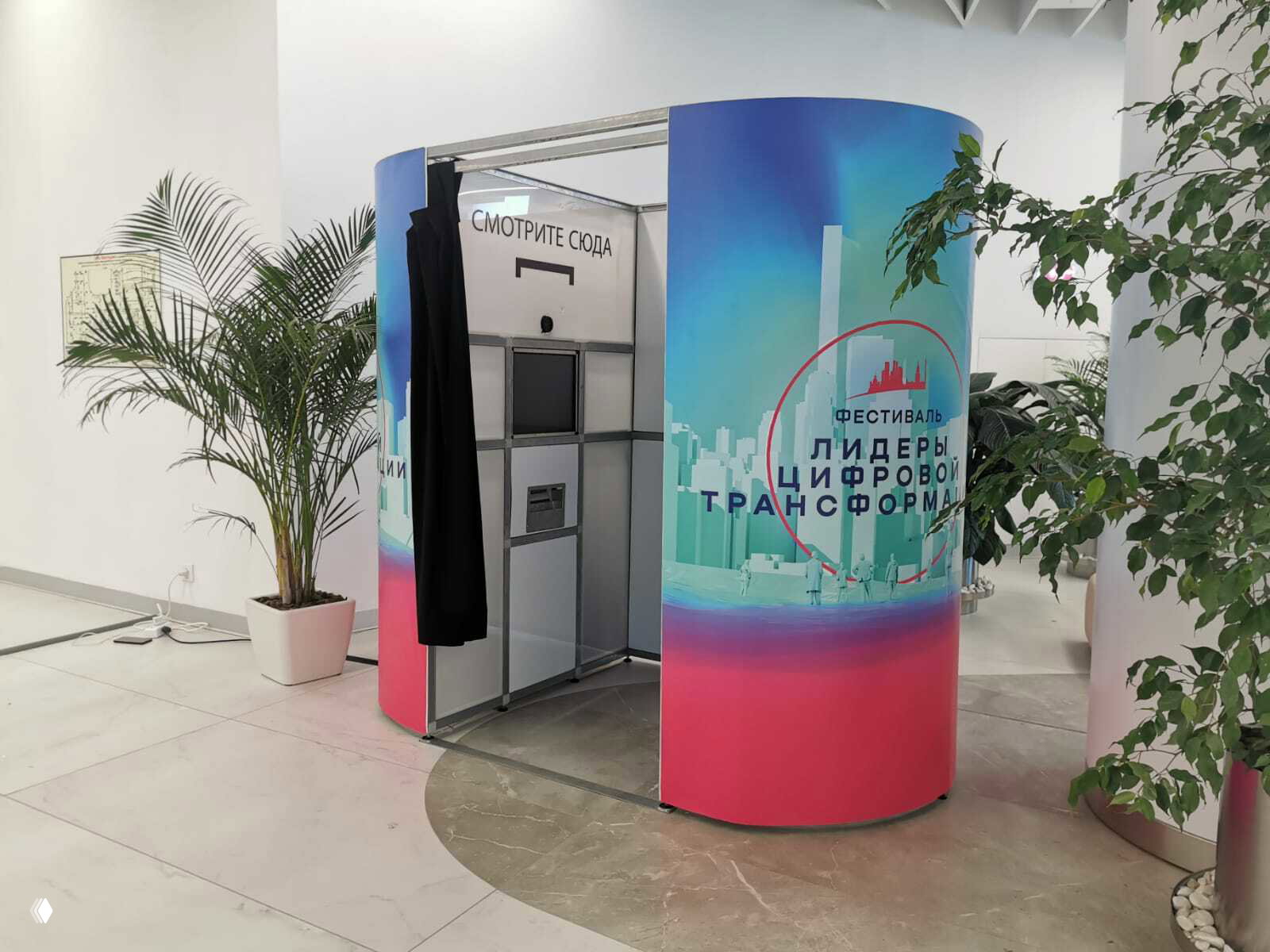 Фотокабина со шторкой под другим углом: видна внутренняя панель с экраном и принтером, крупный логотип мероприятия на внешней обшивке.