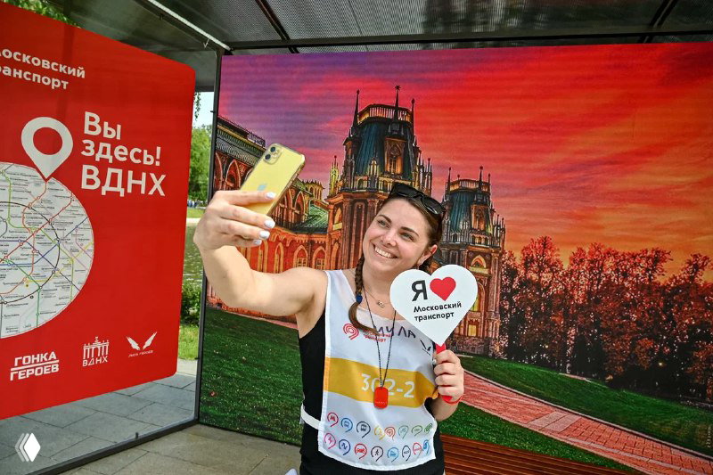 Улыбающаяся девушка делает селфи на фоне светодиодного экрана и указателя «Вы здесь! ВДНХ» — демонстрация фотозоны с LED-фоном для событий.