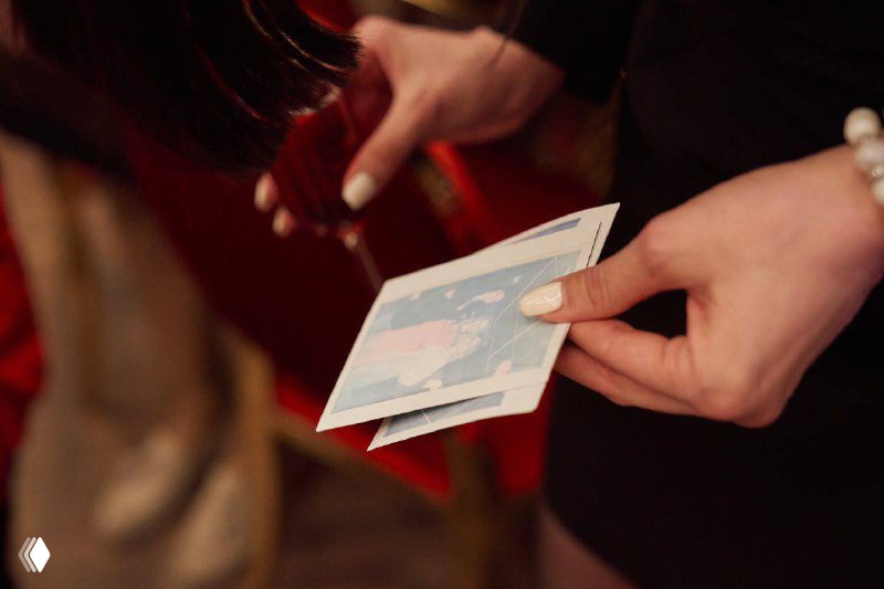 Руки держат только что распечатанную моментальную фотографию формата Instax/Polaroid; видно бумажную фотографию и детали рук гостей на мероприятии.