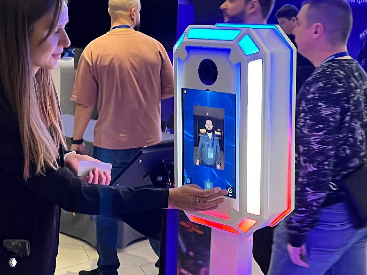 Исходящий кадр пользователя взаимодействующего с белой фотостойкой: рука выбирает опции на сенсорном дисплее аппарата с подсветкой.