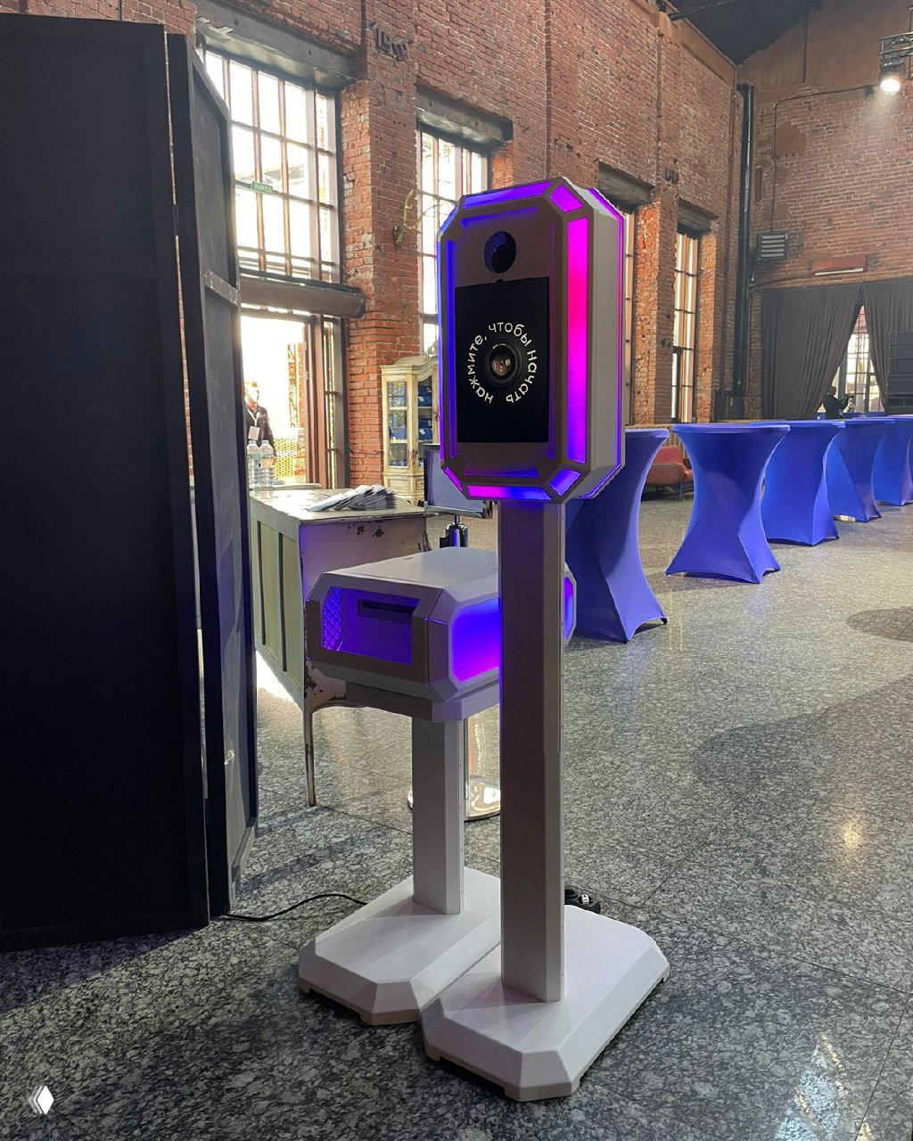 Реальная фотостойка с LED‑подсветкой, установленная в лофтовом зале для мероприятия; видны столы и промышленный интерьер.