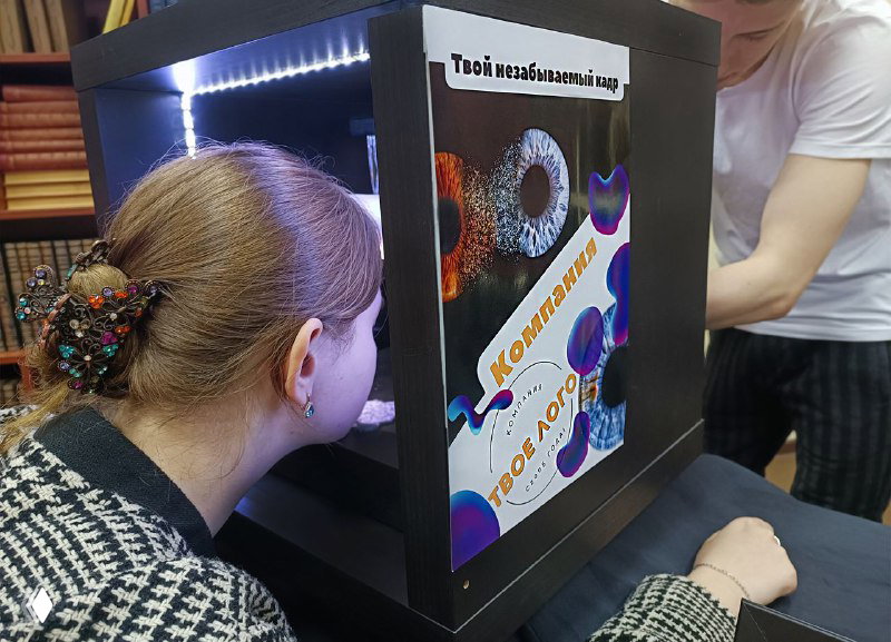 Портативная фотобудка и человек, наклоняющийся к аппарату для съёмки радужки глаза на мероприятии; интерактивная стойка в рабочем окружении