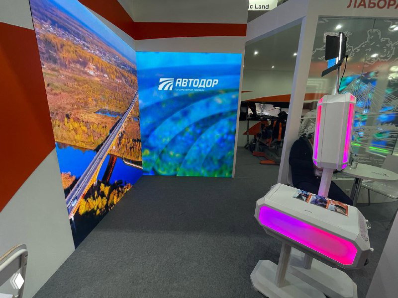 Угол выставочной фотозоны с брендированным изображением на LED‑экране и рядом расположенной белой фотостойкой/GIF‑станцией с подсветкой.