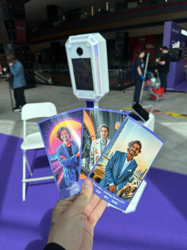 Рука держит напечатанные примеры фотографий с обработкой ИИ на фоне белой фотобудки; демонстрация стилей печати для гостей.